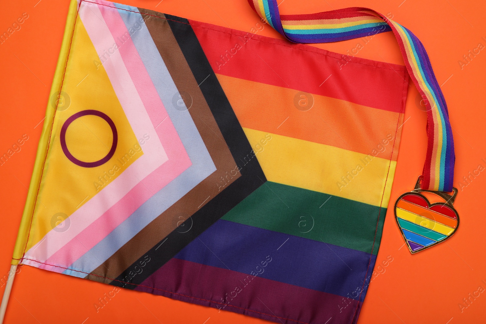 Photo of Bright progress flag and rainbow ribbon with heart shaped pendant on orange background, flat lay. LGBT pride