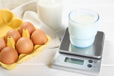Photo of Kitchen scale with glass of milk near eggs on white wooden table, closeup