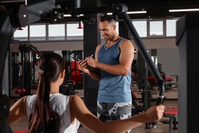 Happy trainer writing down plan of workouts while woman doing exercise in modern gym