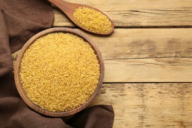 Photo of Bowl and spoon with uncooked bulgur on wooden table, flat lay. Space for text