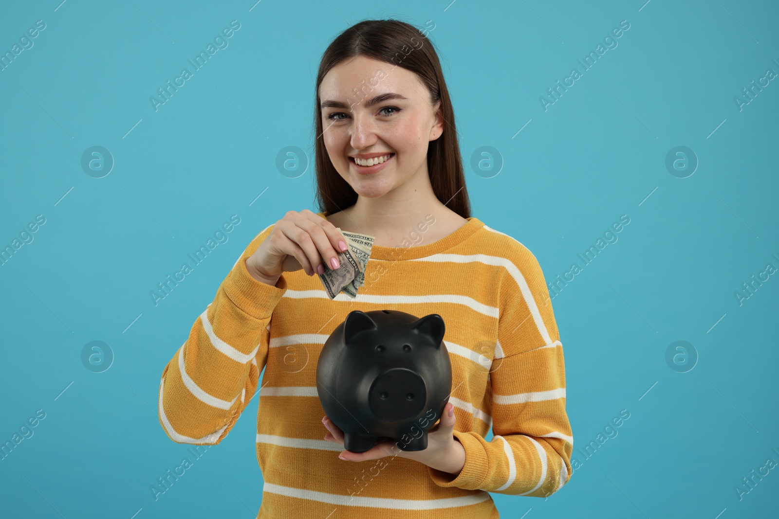 Photo of Happy woman putting dollar banknotes into piggy bank on light blue background