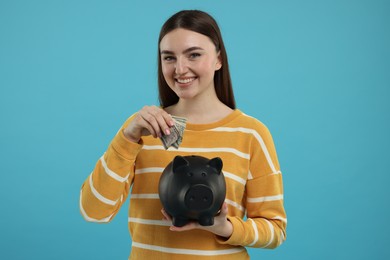 Happy woman putting dollar banknotes into piggy bank on light blue background