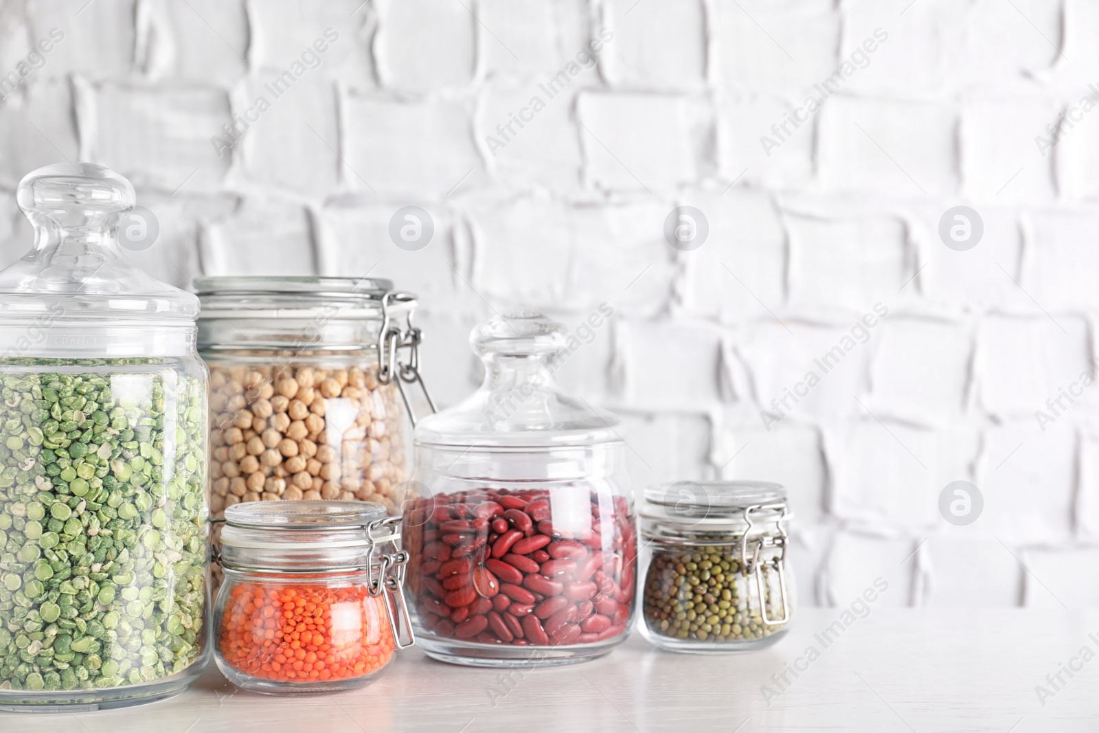 Photo of Jars with different cereals on white table against brick wall