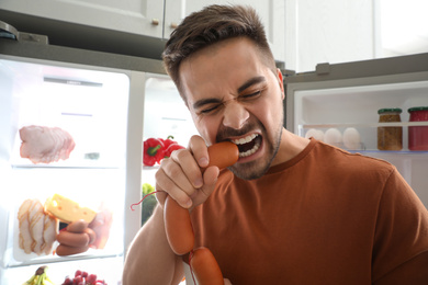 Young man eating sausages near open refrigerator indoors