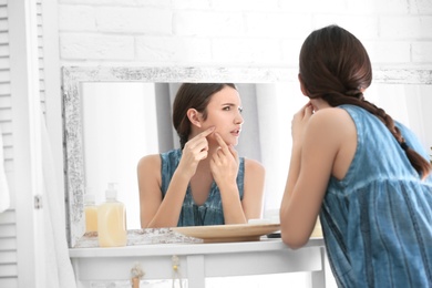 Photo of Teenage girl with acne problem looking in mirror indoors