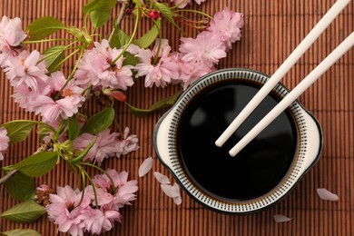 Photo of Bowl of soy sauce, chopsticks and beautiful blossoming branch on bamboo mat, flat lay