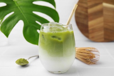 Glass of tasty iced matcha latte, leaf, bamboo whisk and powder on white tiled table, closeup