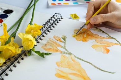 Photo of Woman painting freesias in sketchbook at white table, closeup