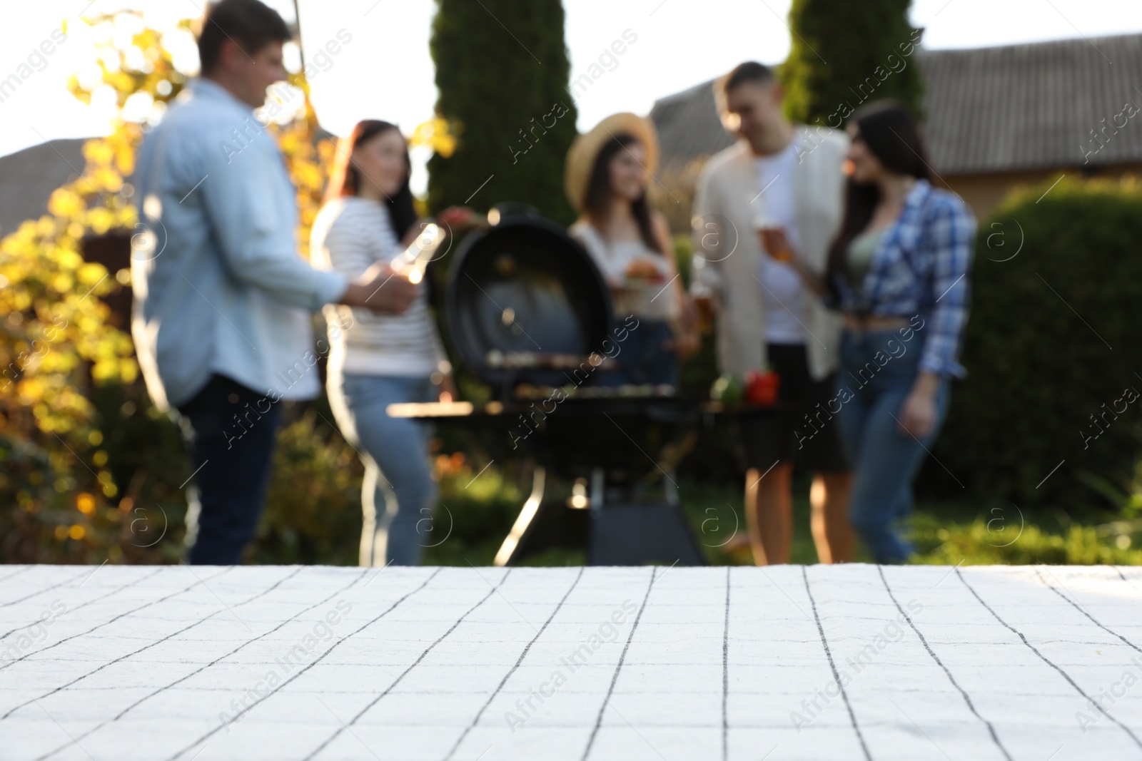 Photo of Empty white wooden table and blurred view of friends having barbecue party outdoors. Space for text