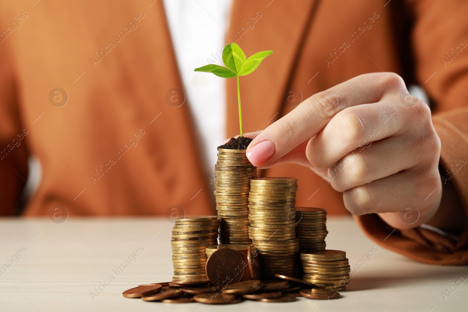 Photo of Woman putting coin with green sprout onto stack at white table, closeup. Investment concept