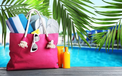 Image of Bag with different beach accessories on grey wooden surface and green palm leaves near outdoor swimming pool