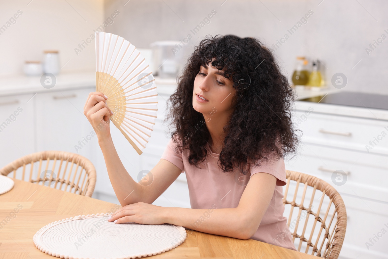 Photo of Young woman waving hand fan to cool herself at table in kitchen