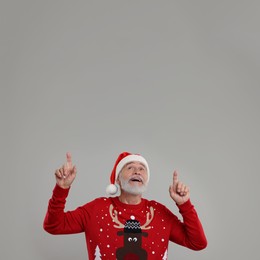 Photo of Senior man in Christmas sweater and Santa hat pointing at something on grey background. Space for text