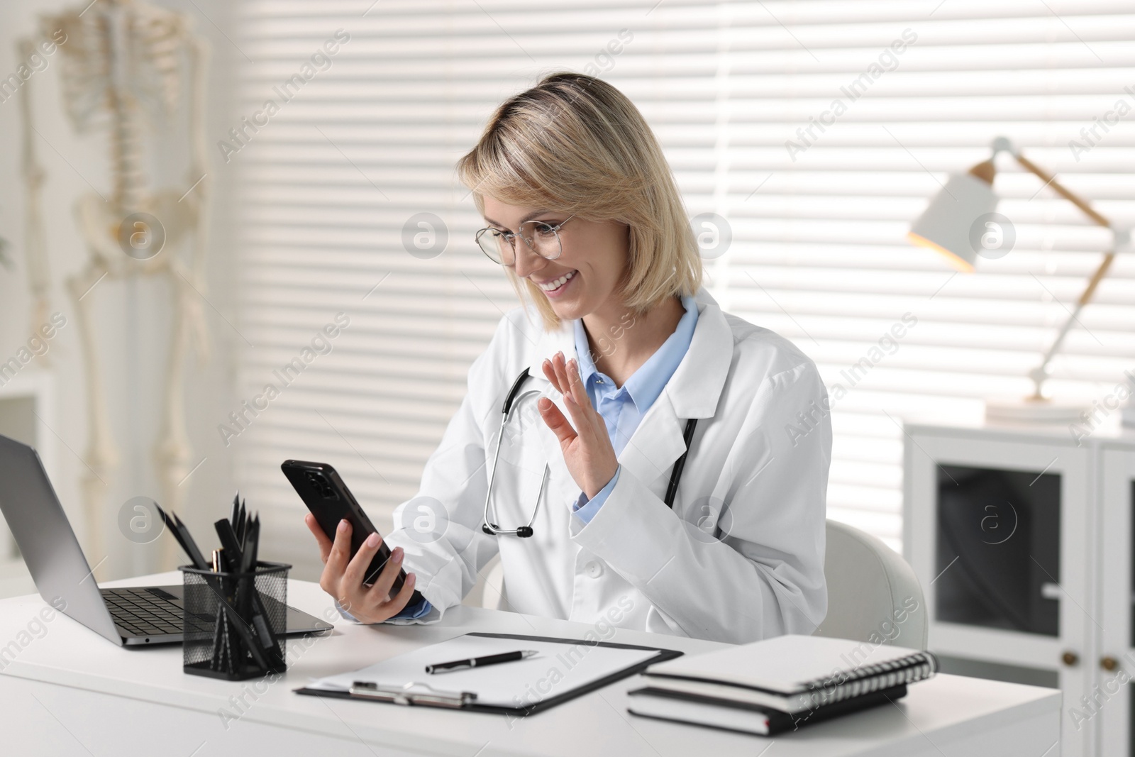 Photo of Smiling doctor with smartphone having online consultation at table in office