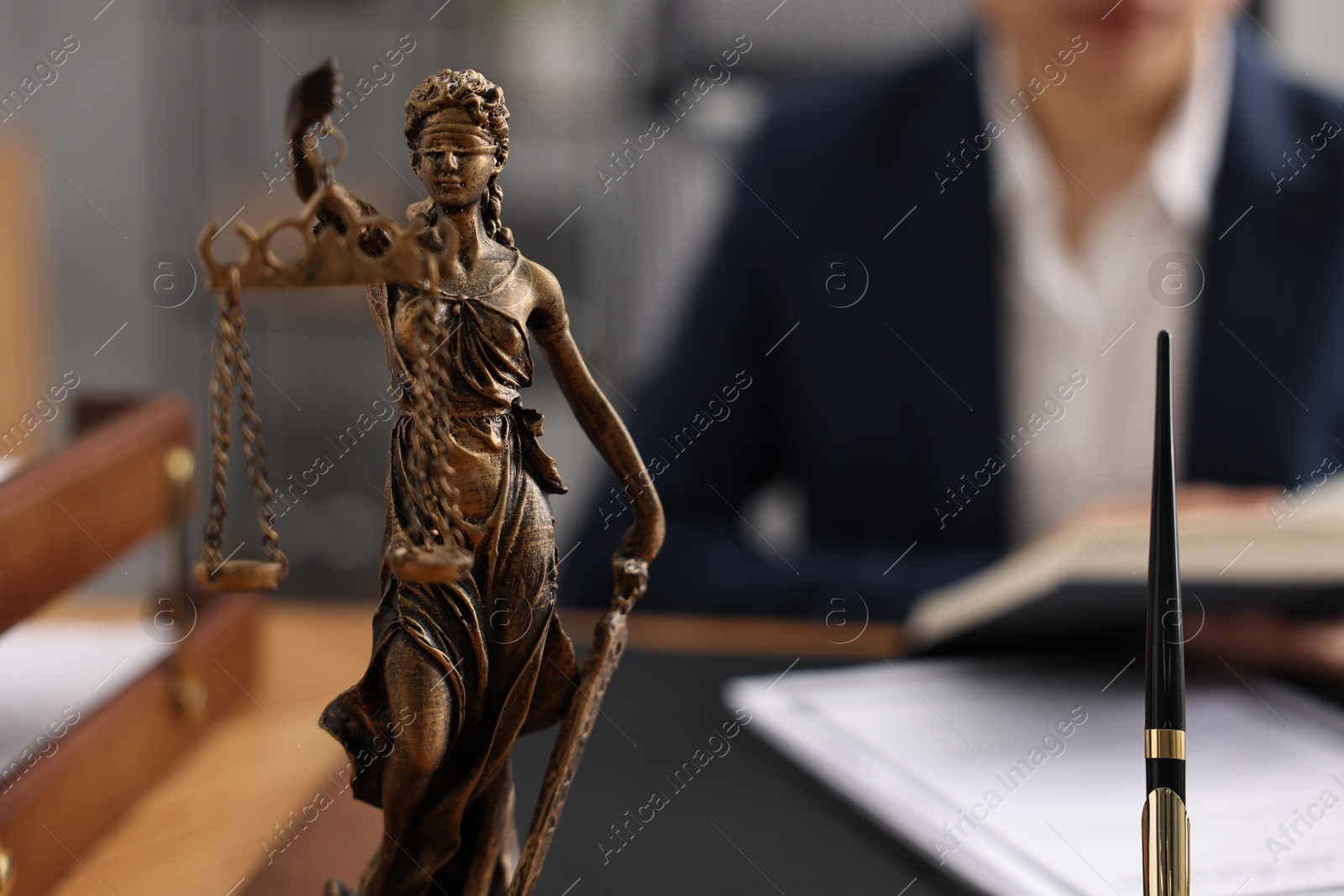 Photo of Notary reading book at table in office, focus on statue of Lady Justice and pen