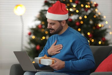 Photo of Celebrating Christmas online with exchanged by mail presents. Man thanking for gift during video call on laptop at home