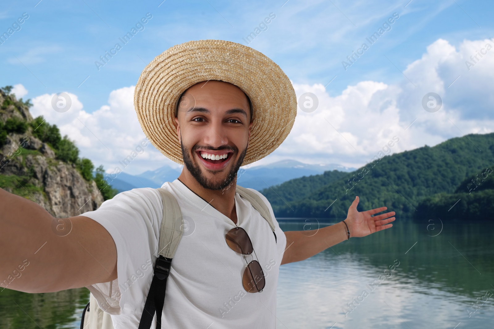 Image of Smiling young man in straw hat taking selfie near river in mountains