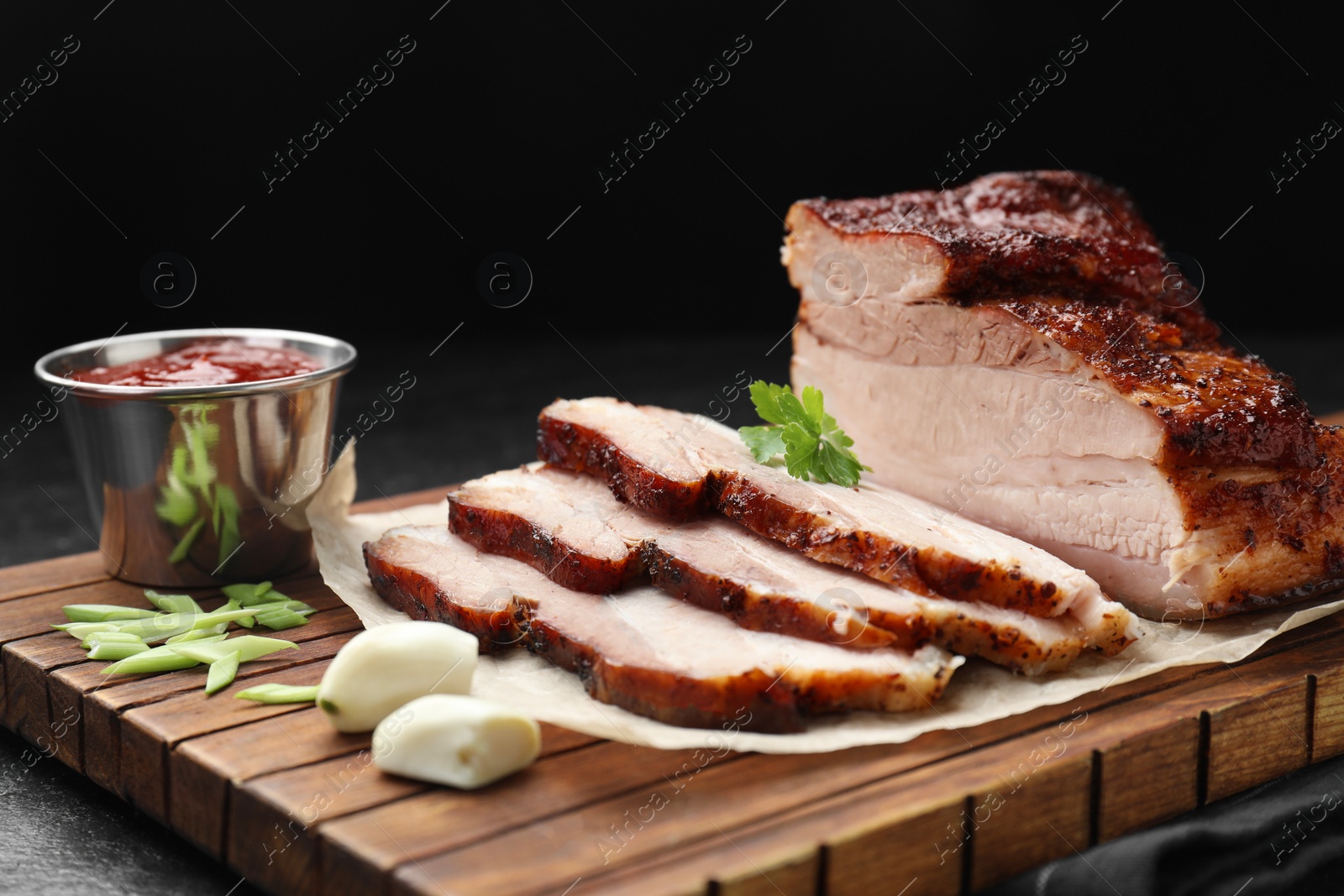 Photo of Pieces of baked pork belly served with sauce and parsley on black textured table, closeup