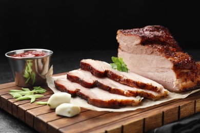 Photo of Pieces of baked pork belly served with sauce and parsley on black textured table, closeup