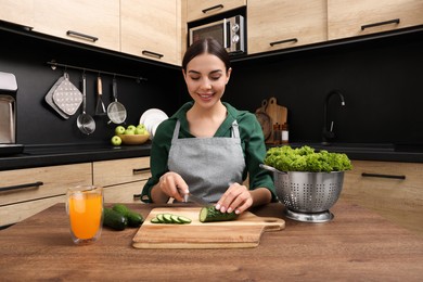 Photo of Woman in apron cutting cucumber at wooden table indoors