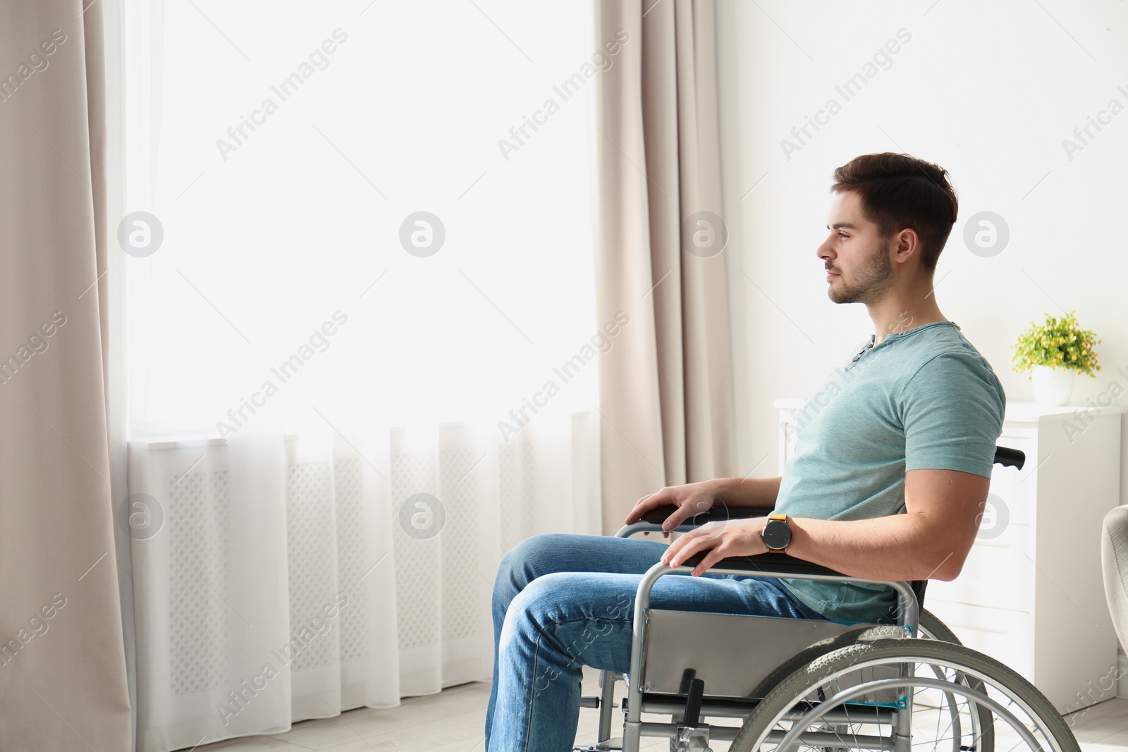 Photo of Young man sitting in wheelchair near window indoors. Space for text