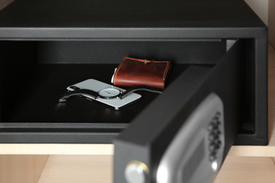 Photo of Open steel safe with wallet, watch and smartphone in wooden closet at hotel