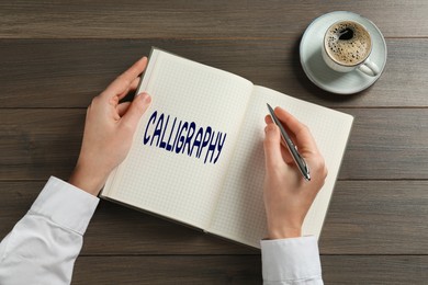 Woman writing word Calligraphy in notebook at wooden table, top view