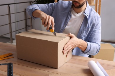 Photo of Man using utility knife to open parcel at wooden table indoors, closeup