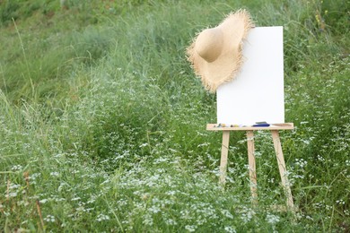 Photo of Wooden easel with blank canvas, painting equipment and hat in meadow. Space for text