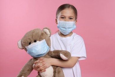 Photo of Cute girl and teddy bear with medical masks on pink background