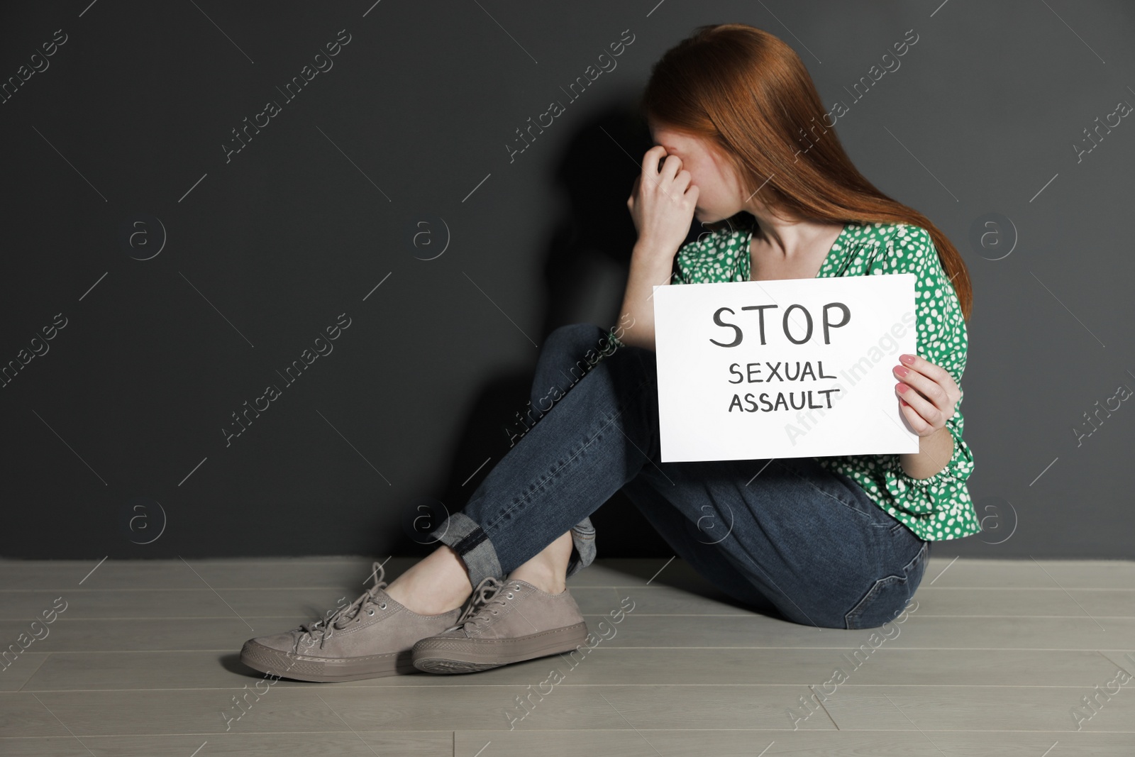 Photo of Young woman holding card with words STOP SEXUAL ASSAULT while sitting near grey wall. Space for text