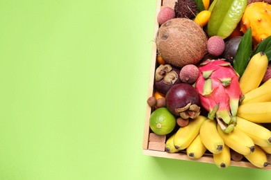 Photo of Different tropical fruits in wooden box on green background, top view. Space for text