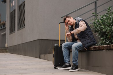 Photo of Being late. Worried man with suitcase looking at watch on bench outdoors, space for text