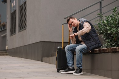 Being late. Worried man with suitcase looking at watch on bench outdoors, space for text