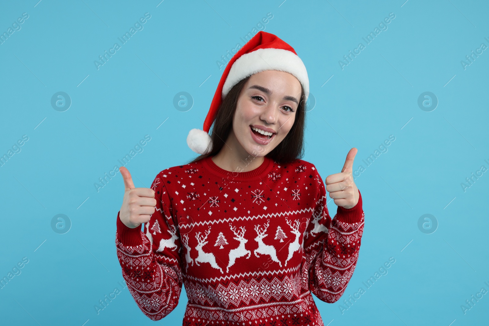 Photo of Happy young woman in Christmas sweater and Santa hat showing thumbs up on light blue background
