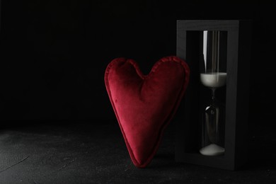 Photo of Heart shaped cushion and hourglass on black stone table, space for text. Relationship problems concept