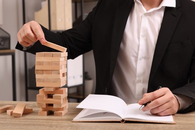 Playing Jenga. Businessman building tower with wooden blocks and writing result into notebook at table indoors, closeup