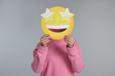 Woman holding emoticon with stars instead of eyes on grey background
