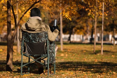 Photo of Man with thermos sitting in camping chair outdoors on autumn sunny day, back view
