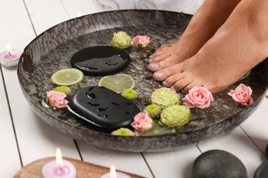 Photo of Woman soaking her feet in plate with water, stones, flowers and lime slices on white wooden floor, closeup. Pedicure procedure