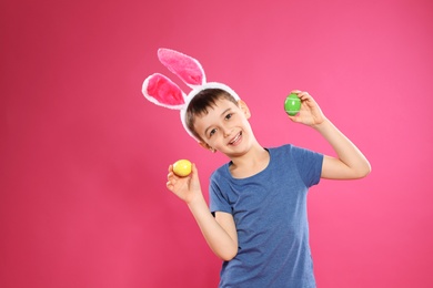 Little boy in bunny ears headband holding Easter eggs on color background, space for text