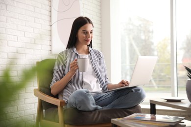 Photo of Beautiful young woman with cup of drink using laptop at home