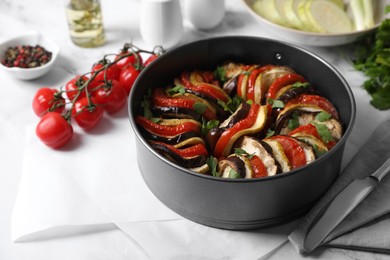 Photo of Delicious ratatouille in round baking pan, knife and ingredients on white table