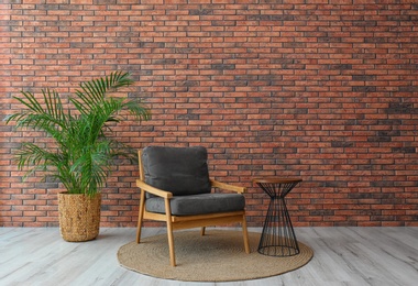 Modern room interior with stylish grey armchair and potted plant near brick wall