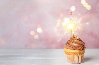 Image of Birthday cupcake with sparkler on white wooden table against light pink background. Space for text