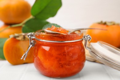 Photo of Jar and spoon of tasty persimmon jam, ingredients on white tiled table, closeup