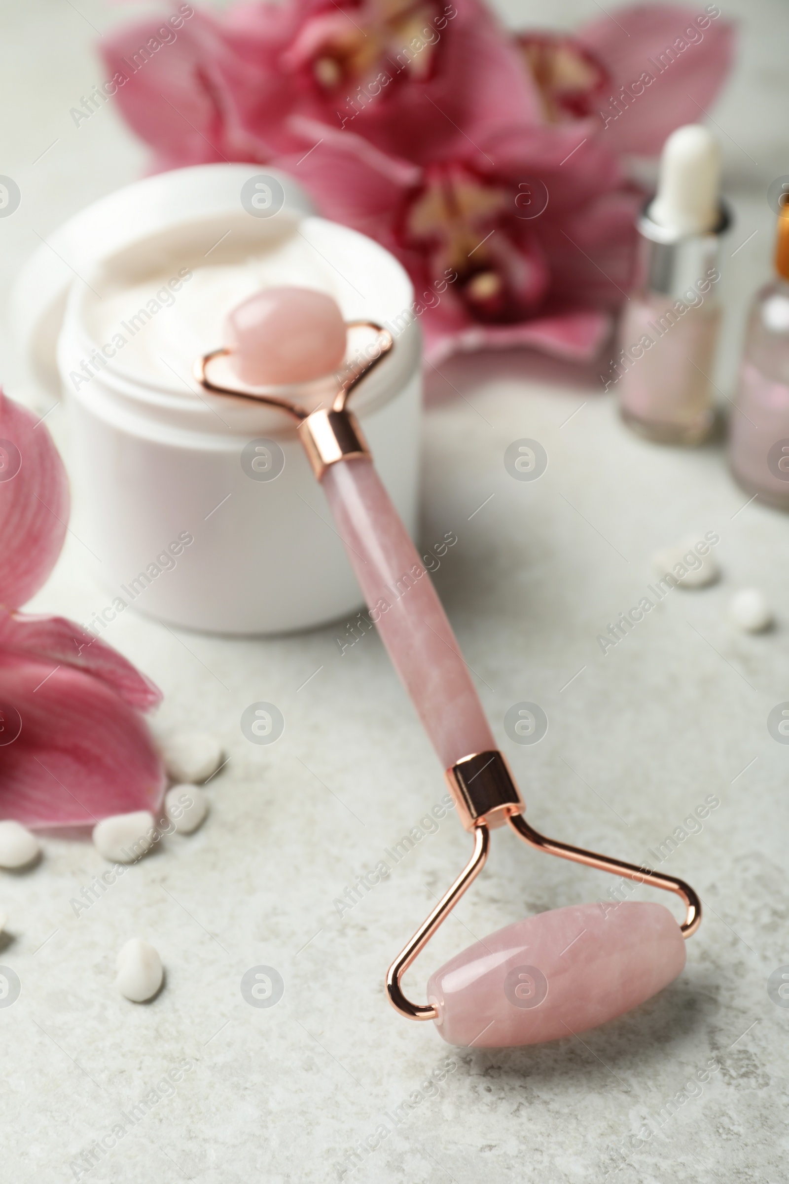 Photo of Natural rose quartz face roller, cosmetic products and flowers on grey background