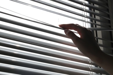 Photo of Woman separating slats of white blinds indoors, closeup