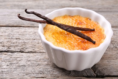 Photo of Delicious creme brulee in bowl and vanilla pods on wooden table, closeup