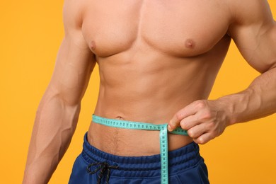 Photo of Athletic man measuring waist with tape on orange background, closeup. Weight loss concept